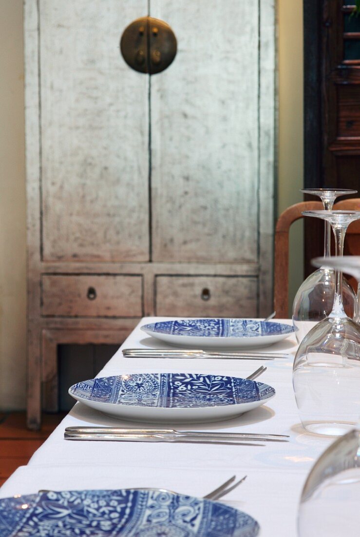 Weiß-blaue Teller und Besteck auf weißem Tischtuch neben asiatischem Holzschrank an Wand