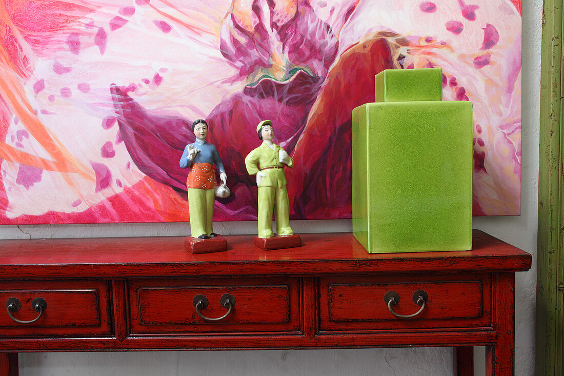 Bemalte Porzellanfiguren neben grünem Keramikbehälter mit Deckel auf Wandtisch und buntes Bild an Wand