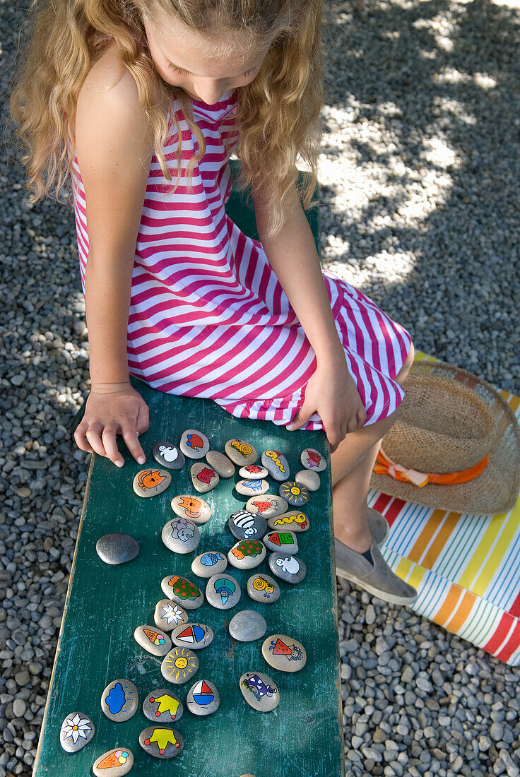 Girl playing with painted stones on wooden bench in gravel garden