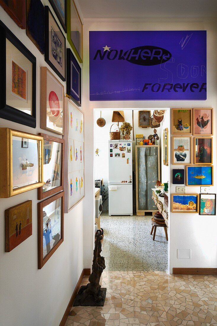 Vorraum mit dichtgehängter Bildergalerie und blauviolettes Schriftplakat über Durchgang zur improvisierten Küche