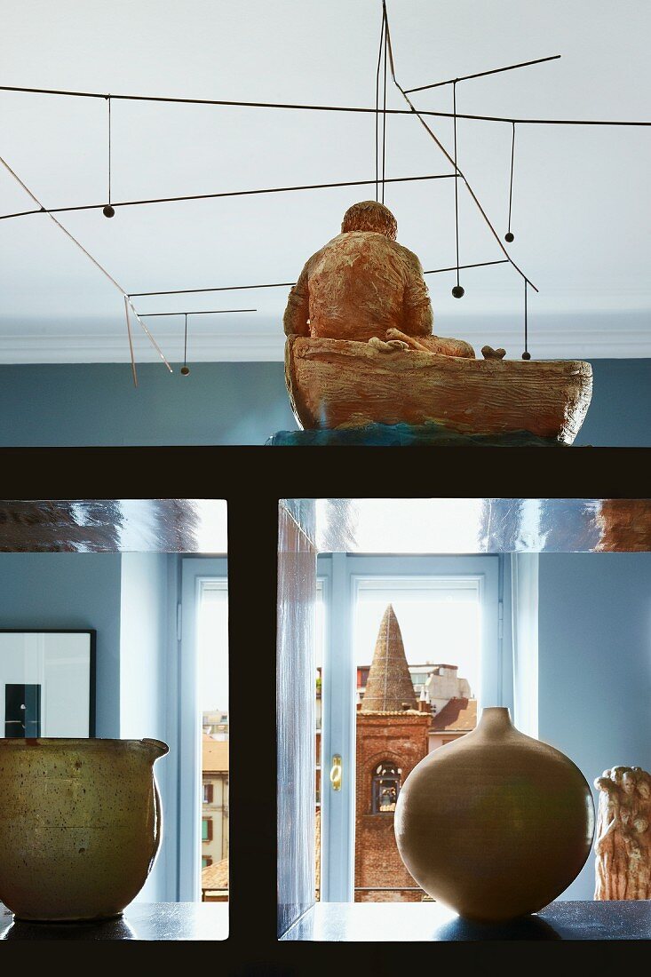 Blick durch Regalraumteiler mit Vasen und figürlicher Skulptur auf dahinterliegende hellblaue Fensterwand und Ausblick auf nahen italienischen Glockenturm