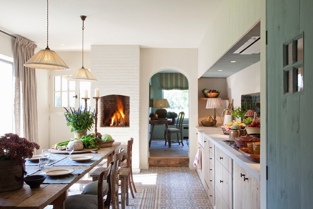 Langer Holztisch mit Gedecken in schlichter Küche mit offenem Kamin und Blick durch Rundbogen auf Tisch und Stuhl
