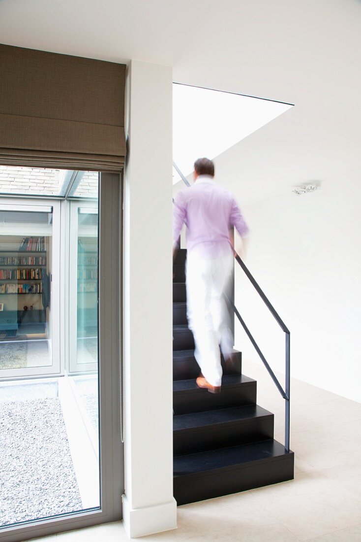 Man walking up black metal staircase in contemporary house with view of courtyard through terrace windows