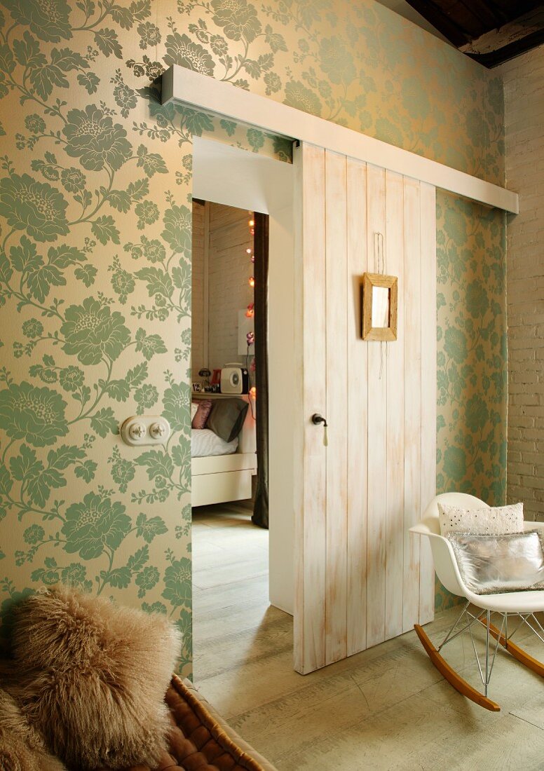 Klassiker Schaukelstuhl vor tapezierter Wand mit elegantem Blumenmuster und halboffener Schiebetür mit Blick in Schlafraum