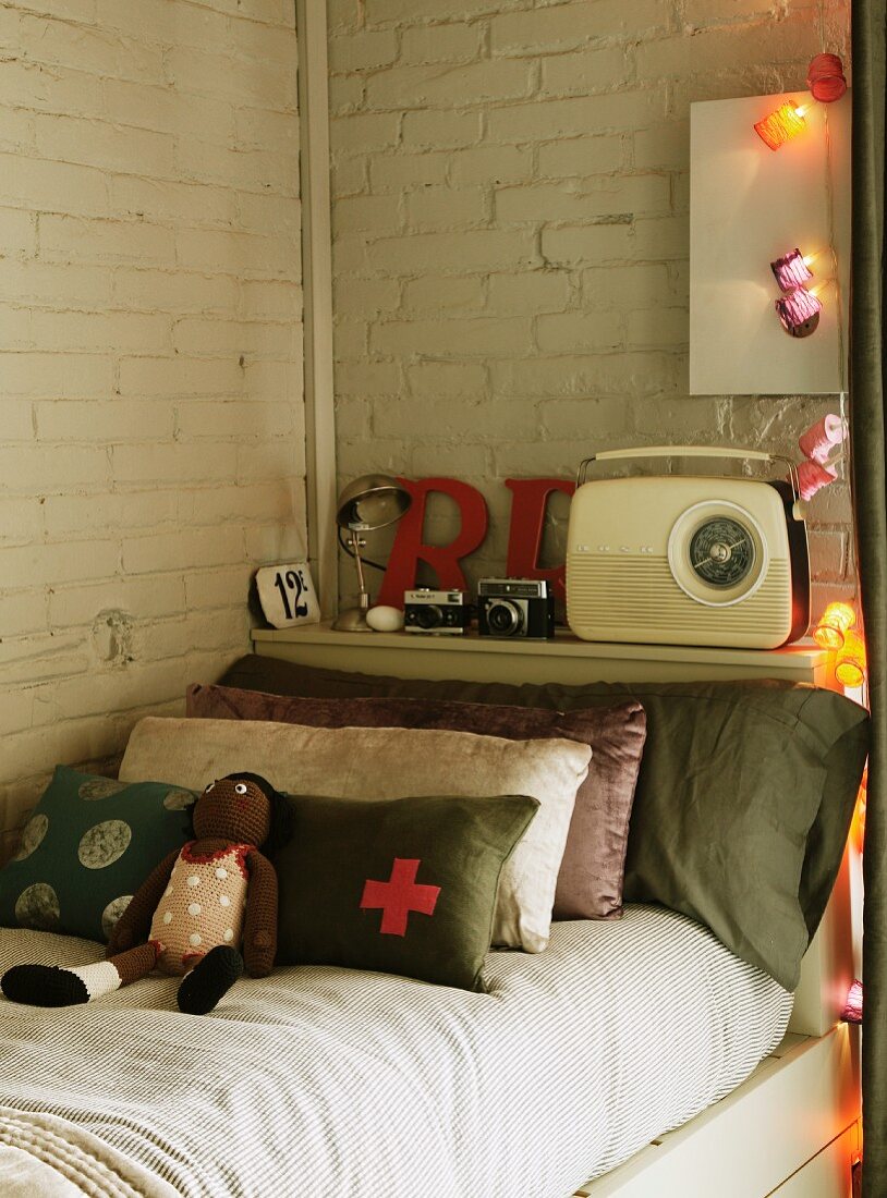 Stoffpuppe und Kissenstapel am Kopfende des Bettes vor Ablage mit Retro Radio an getünchter Ziegelwand