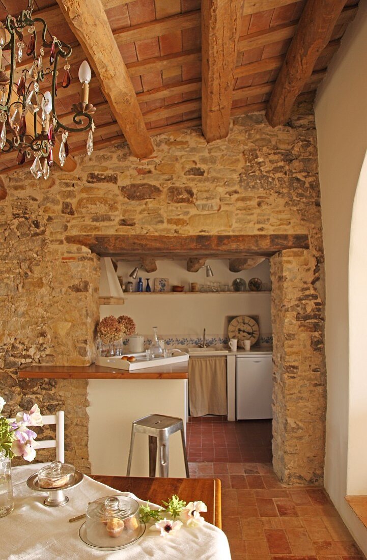 View of stone wall and doorway leading to simple, white kitchen from dining area with Spanish chandelier
