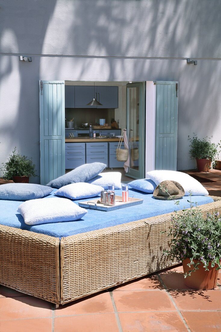 Rattanbett mit blauem Laken und Kissen auf sonniger Terrasse; im Hintergrund Blick in die Küche des modernen, mediterranen Wohnhauses