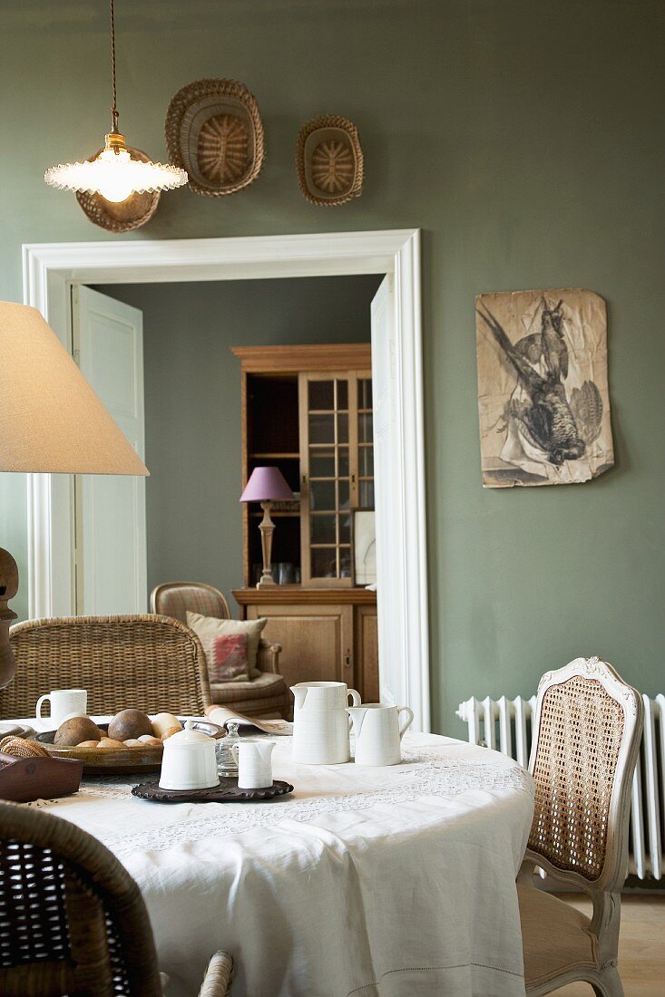 Blick über gedeckten Frühstückstisch mit Korb- und Geflechtstühlen auf blassgrün getönte Wände in Wohn- und Esszimmer