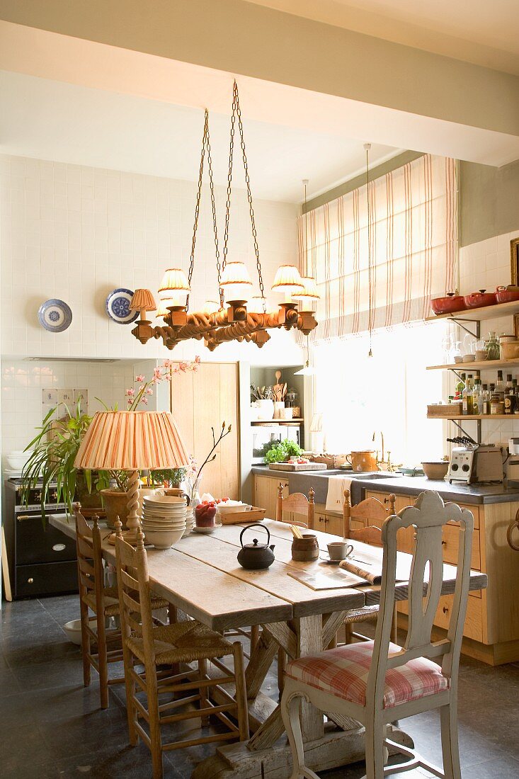 Wooden table and various wooden chairs below rustic pendant lamp in eclectic yet tasteful kitchen-dining room