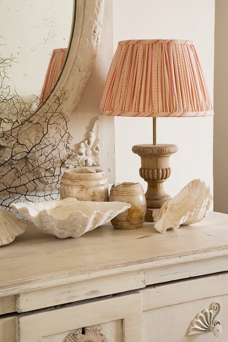 Shabby-chic arrangement of sea shells, marble pots and lamp with fabric lampshade and wooden base on white chest of drawers