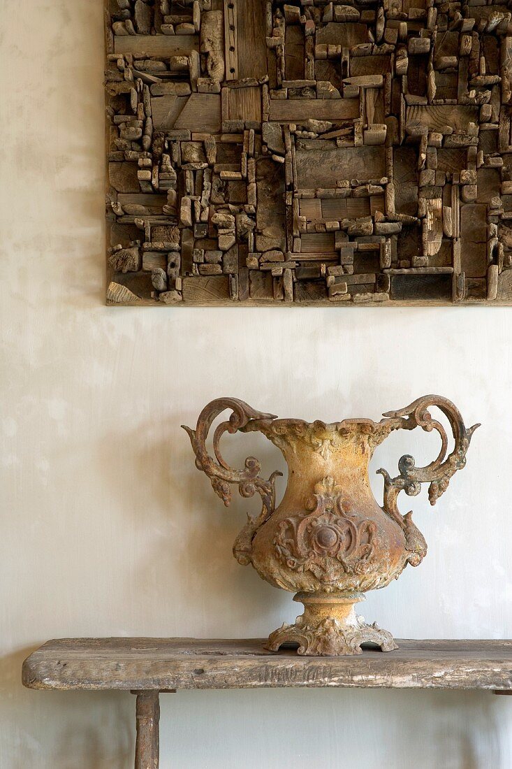 Antike, gusseiserne Vase mit Fuss auf rustikaler Holzbank und Setzkasten-Holzobjekt an Wand