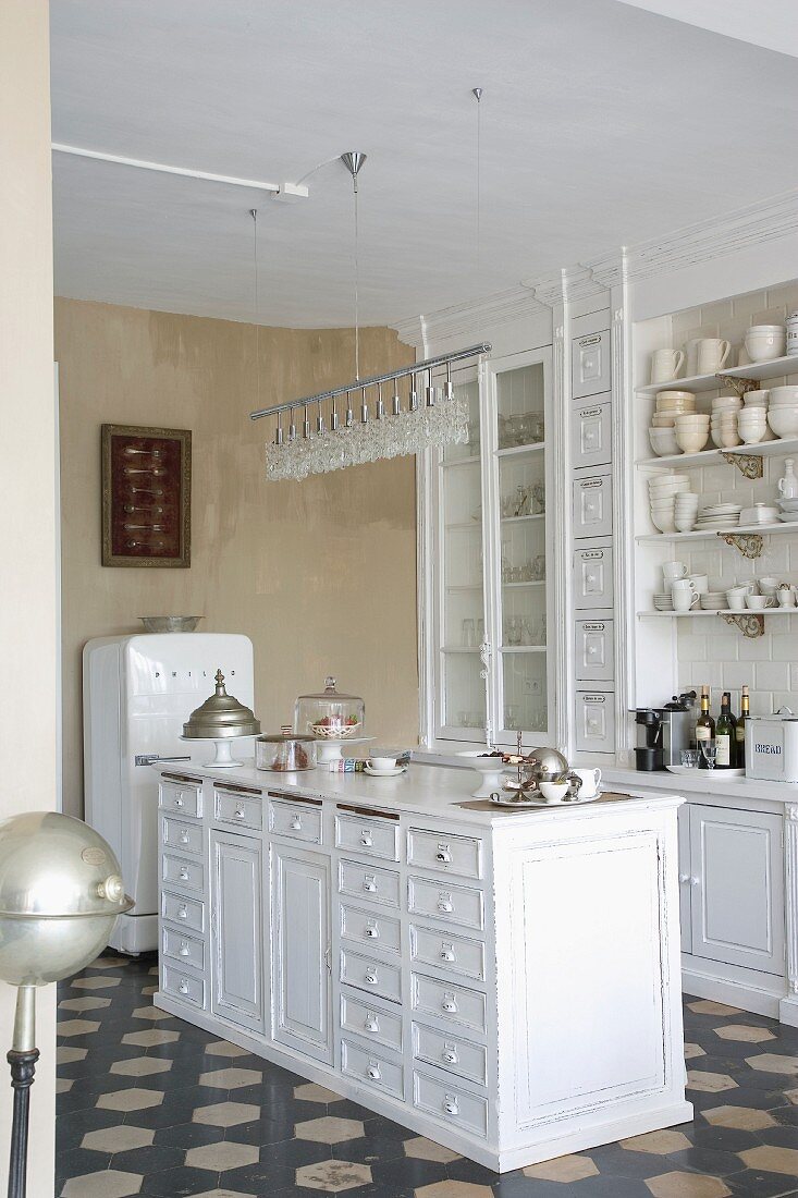 Free-standing, white-painted, country-style wooden counter with drawers below modern pendant lamp and fitted cupboards on vintage tiled floor