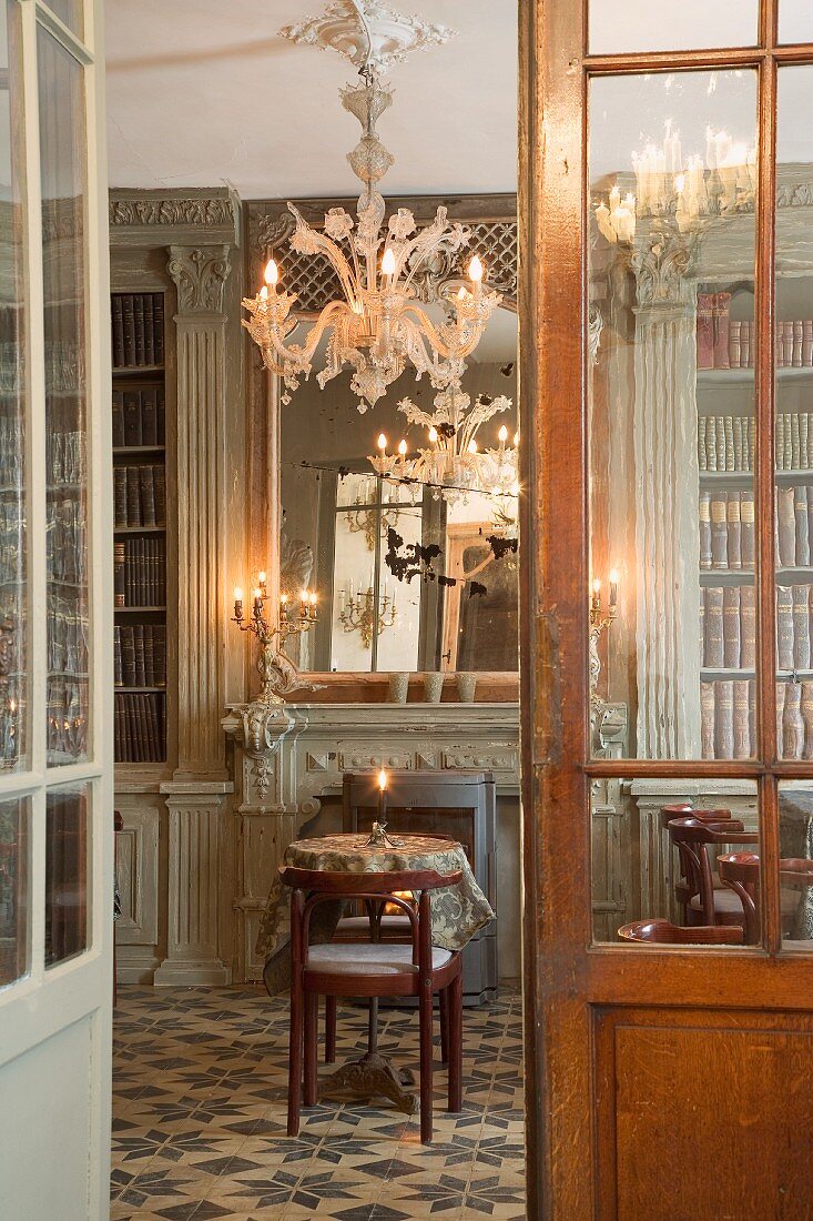 View through open door into library with round table and chair in front of fireplace below floral chandelier