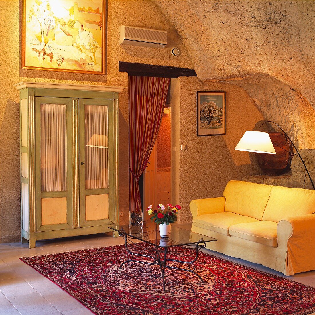 Glastisch auf Orientteppich vor gelber Couch und schlichter Bauern-Vitrinenschrank im Wohnraum mit felsenartiger Wand