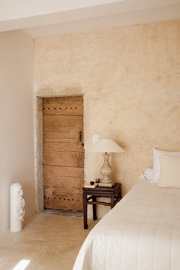Antikes Nachtkästchen mit Tischlampe neben rustikaler Zimmertür in schlichtem Schlafzimmer