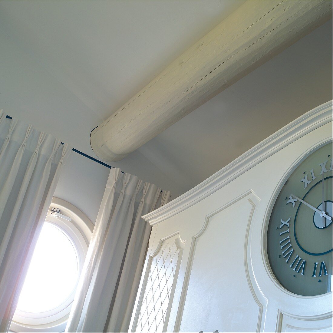 White-painted, traditional cupboard with integrated clock in corner of room next to circular window with curtains
