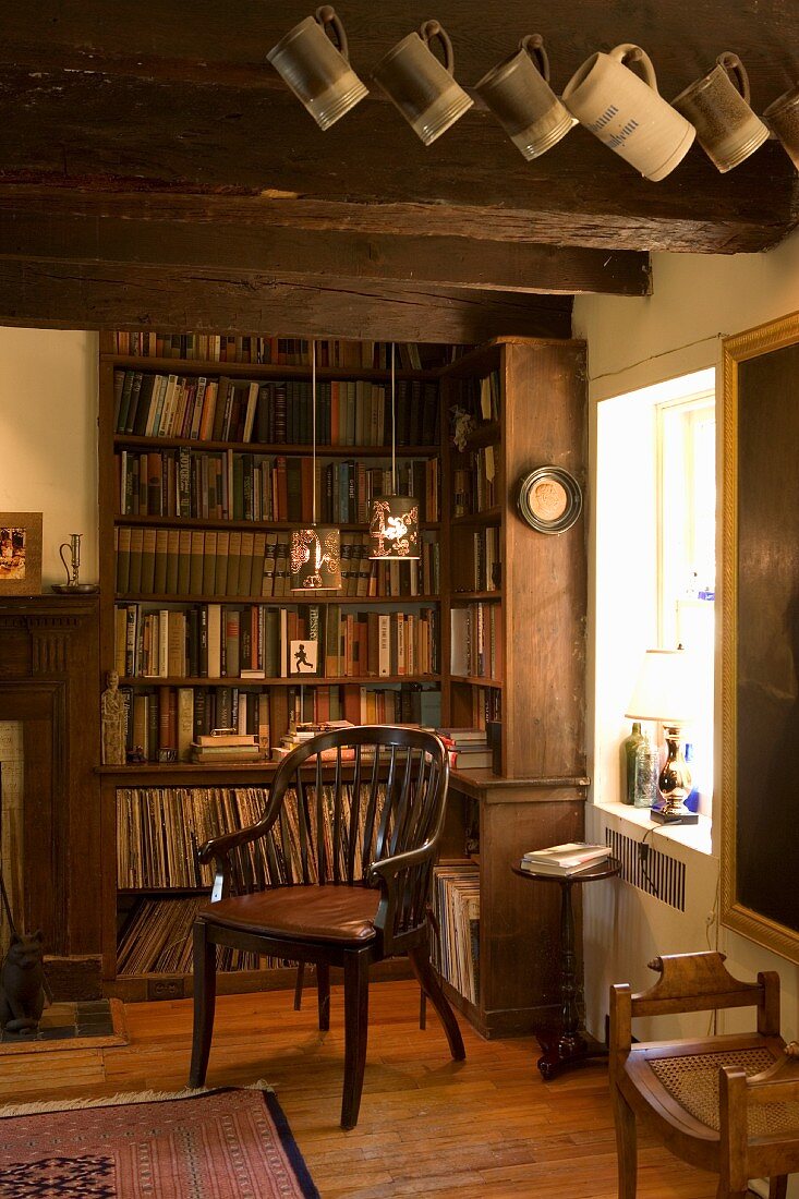 Chair with slatted back in corner of rustic room with fitted bookcase and beer mugs hanging from wooden ceiling beams