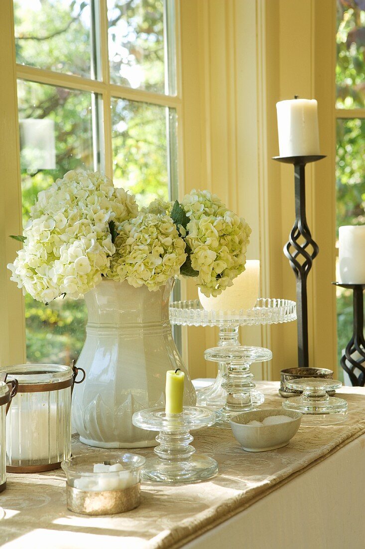 China vase of white hydrangeas and wrought iron candlestick on table below window