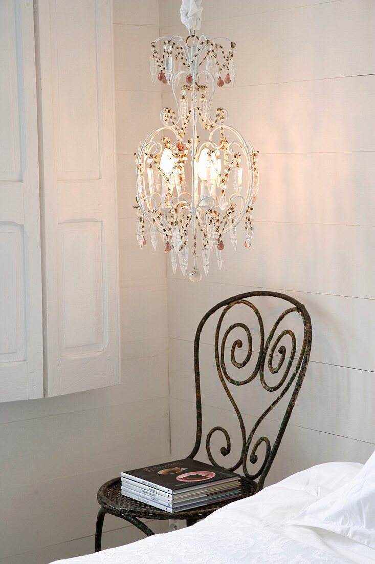 Chandelier with colourful glass pendants and vintage metal chair next to bed against white wooden wall