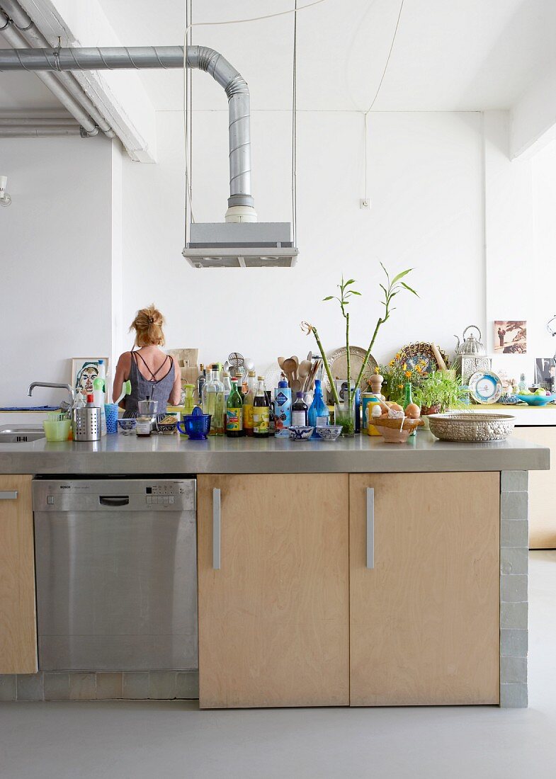 Lebensmitteln und Gewürzutensilien auf freistehender Küchentheke vor Frau im Hintergrund in loftähnlichem Wohnraum