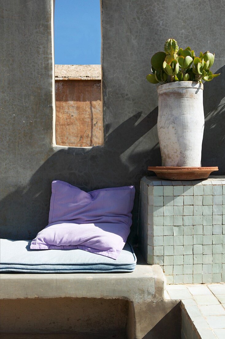 Kissen und Polster auf gemauerter Sitzbank und Pflanzentopf auf Ablage neben fensterartiger Öffnung in Wand
