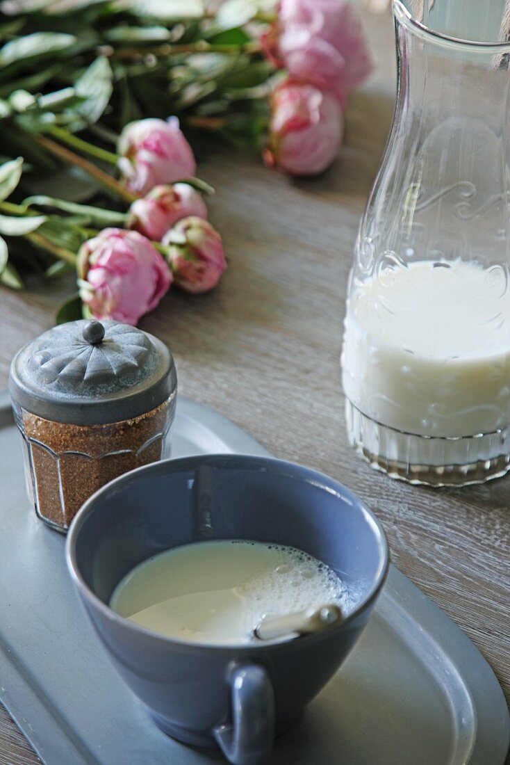 Cup and glass carafe of milk, spice jar and peonies on wooden table