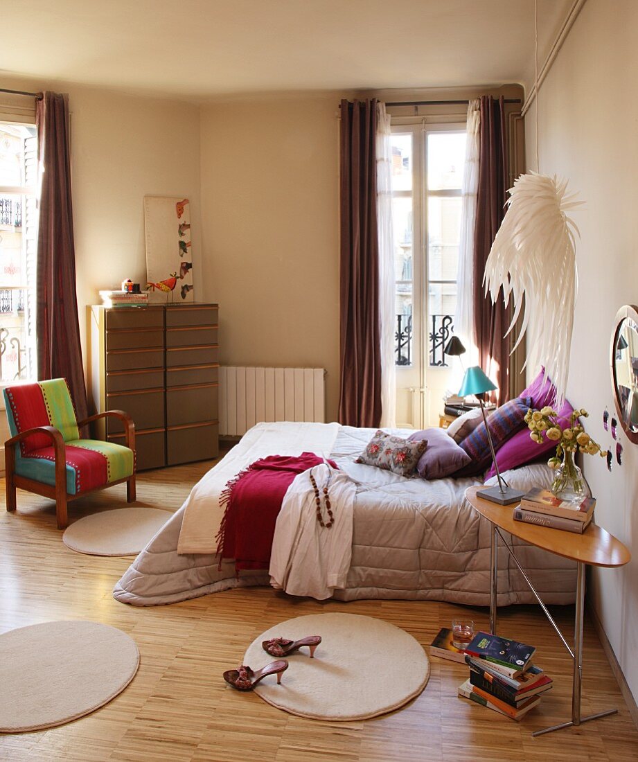 Round rugs around double bed with silver bedspread and vintage chair with colourful upholstery in front of French windows