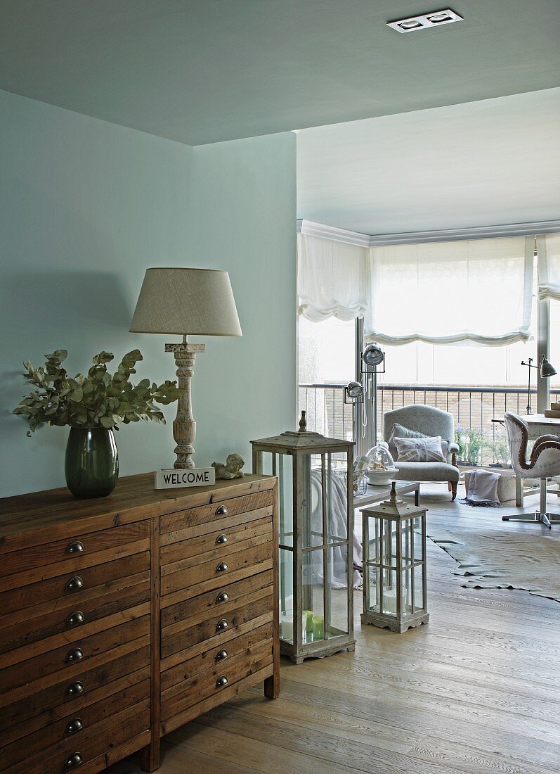 Halbhoher Vintage Schubladenschrank aus Holz an Wand in offenem Wohnzimmer mit Blick auf Sessel vor Fenster und halboffenem Raffrollo