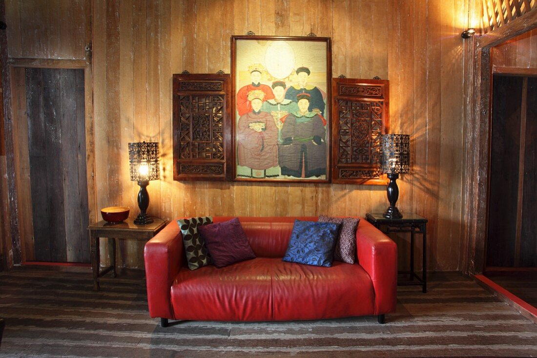 Rote Ledercouch zwischen Beistelltischen an schlichter Holzwand unter Familienbild und geschnitzten Holzschränkchen