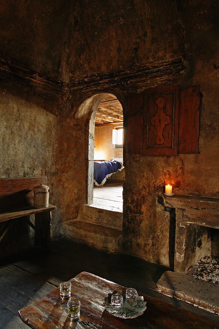 Kaminzimmer in altem Haus mit verrussten Wänden und Blick durch offenen Durchgang mit Rundbogen auf ein Bett