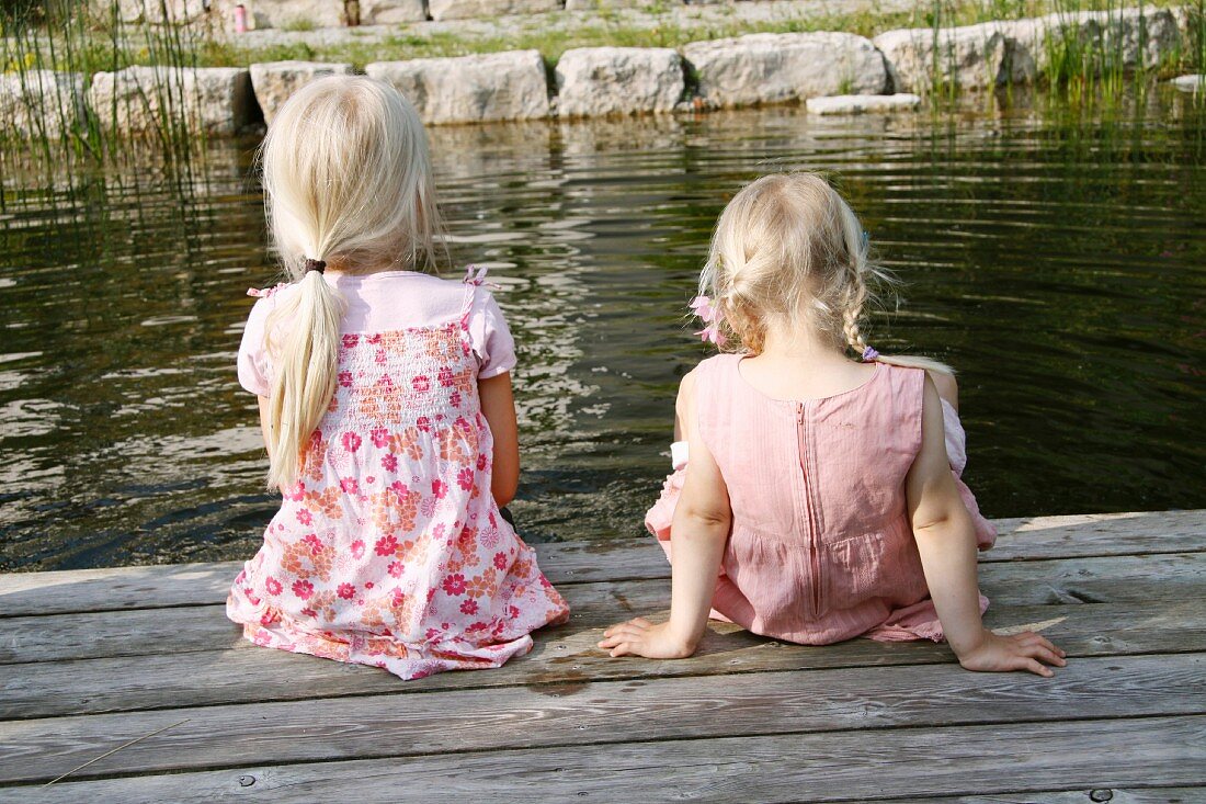 Two blonde girls sitting next to a garden pond
