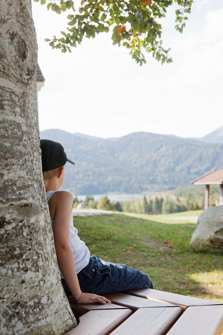 Junge sitzt auf Holzbank unter einem Baum