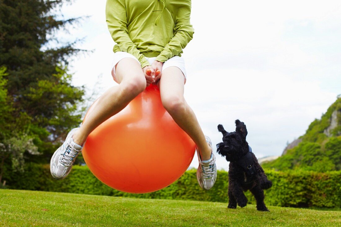 Frau auf einem Hüpfball spielt mit Hund im Garten