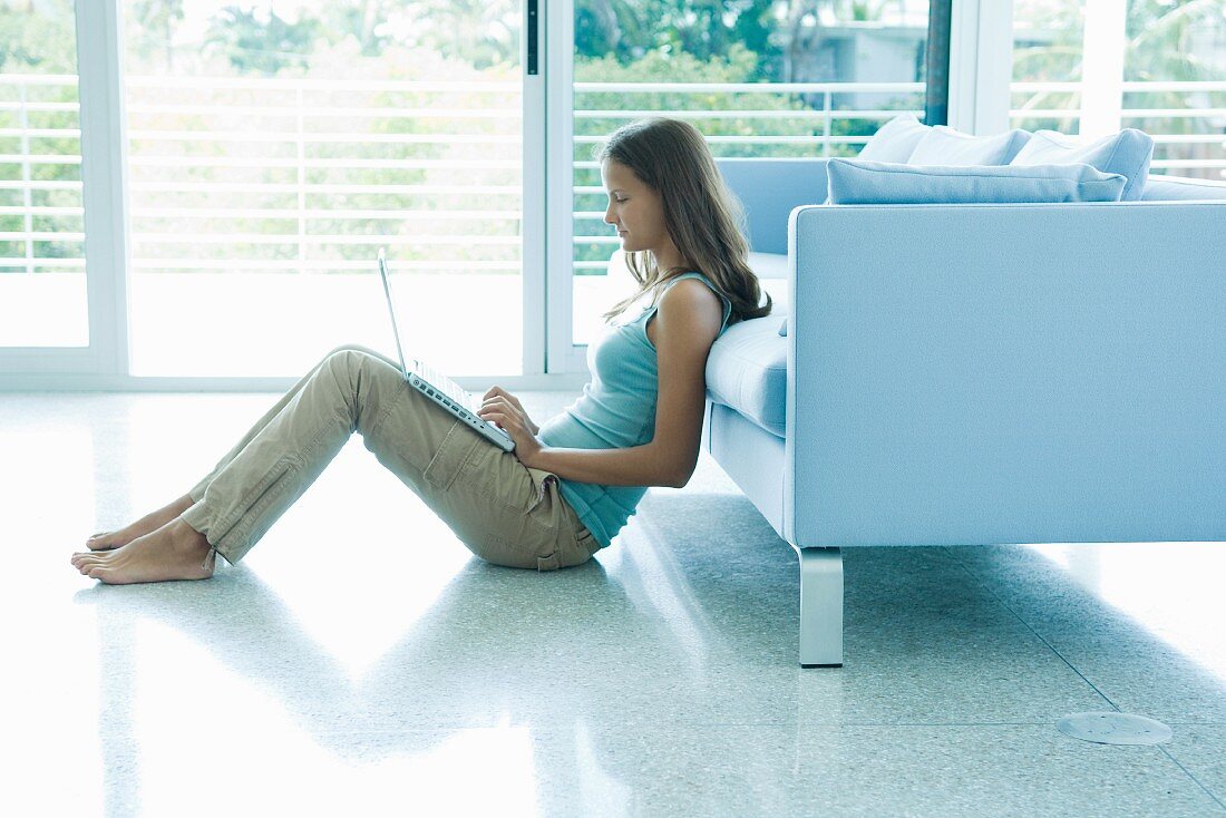 Teenage girl leaning against sofa, using laptop computer, full length