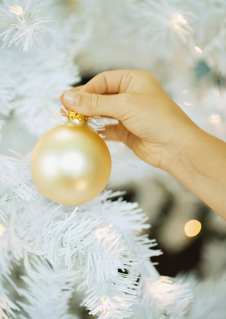 Goldene Weihnachtskugel in der Hand haltend vor künstlichem Weihnachtsbaum