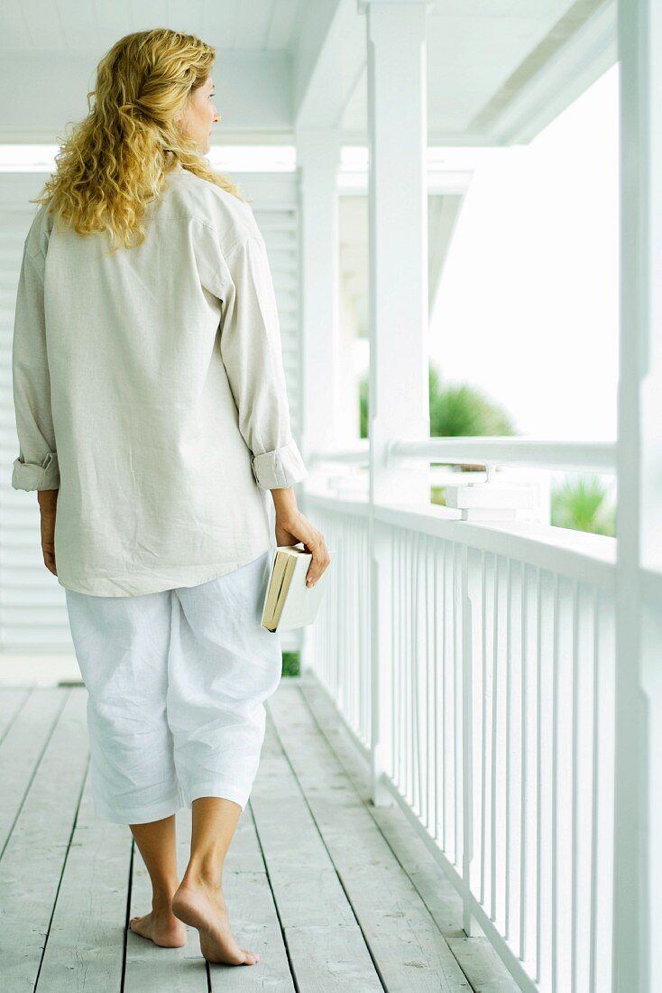 Woman walking on porch, carrying book, rear view