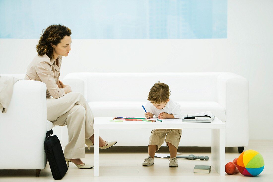 Young professional woman with briefcase sitting in waiting room while son colors