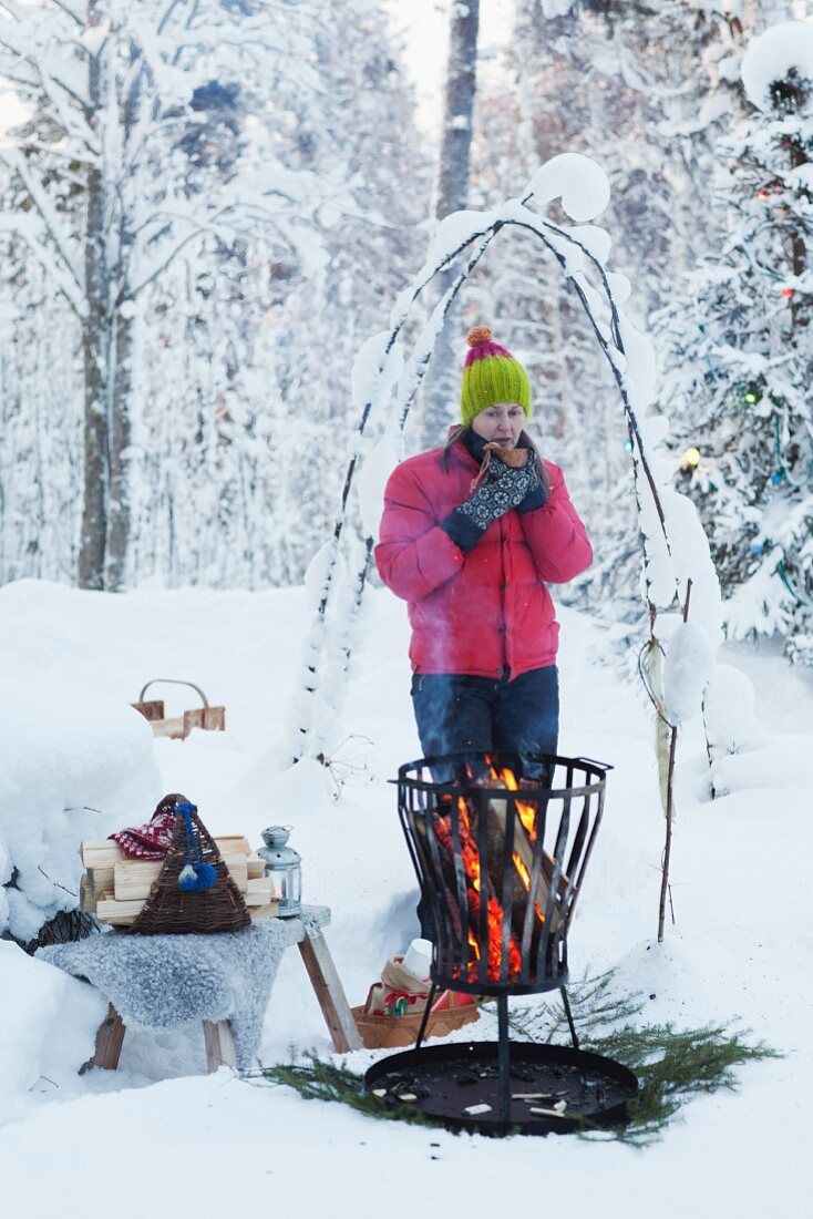 Woman drinking coffee next to fire basket in snowy garden