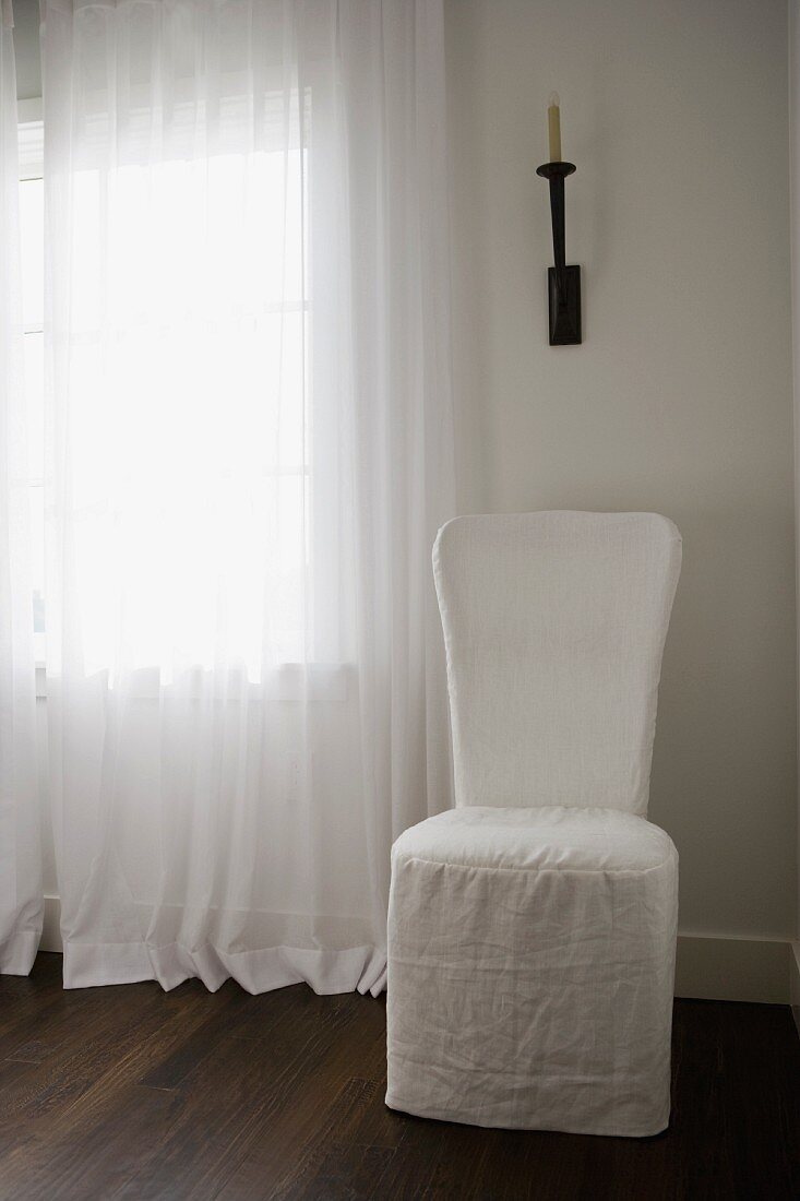 Schlichter Landhausstil - Hussen-Stuhl unter schmiedeeisernem Kerzenleuchter und reinweisser Vorhang auf dunklem Parkett