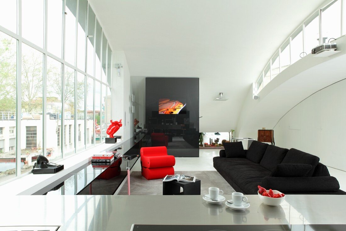 Blick über Edelstahl Küchentheke in offenen Wohnraum mit schwarzem Sofa und rotem Designersessel vor Fensterfront