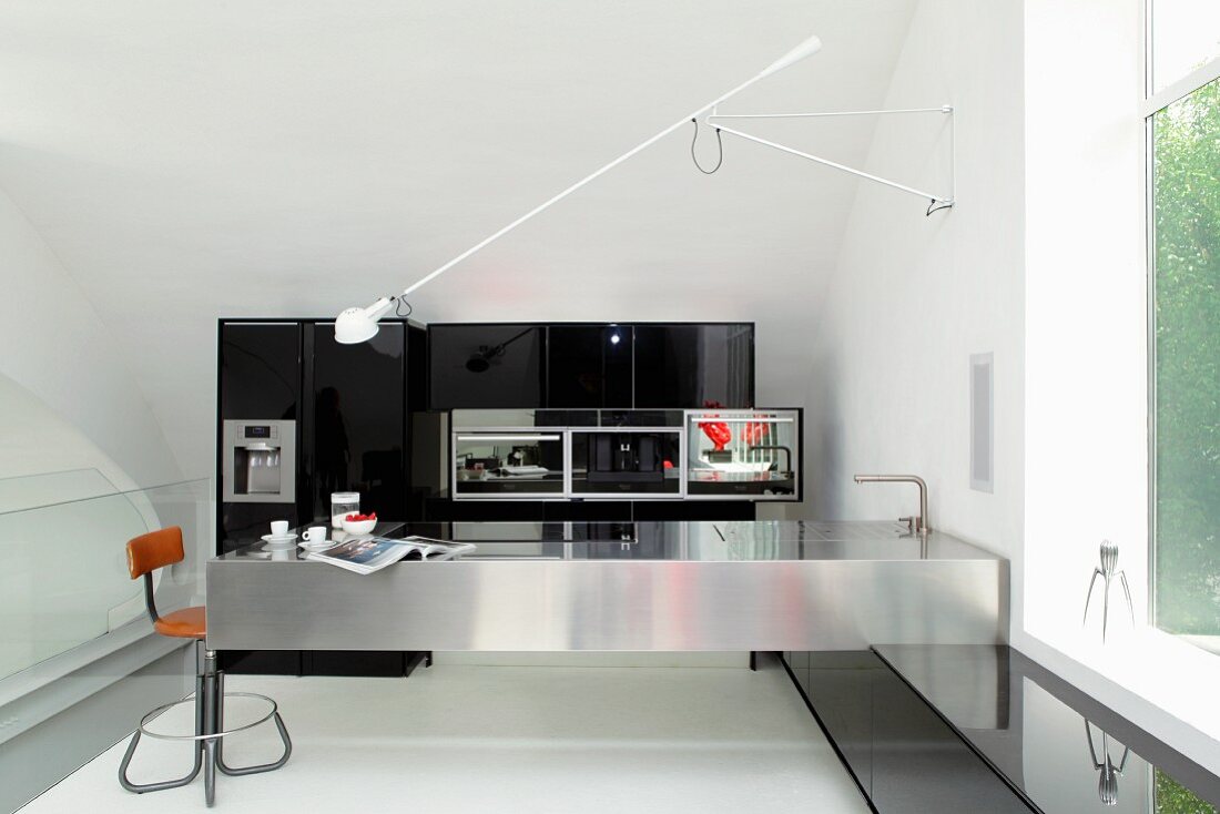 Schwenkarm einer Wandleuchte über Küchentheke aus Edelstahl und Retro Barhocker in minimalistischer Küche mit schwarzem Einbauschrank