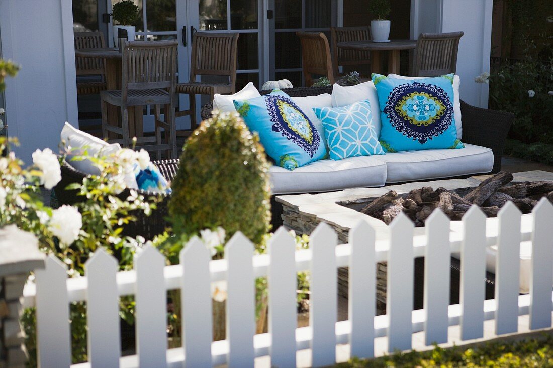Blick über weissen Zaun auf Sitzbank mit bunten Kissen und hellen Polstern auf der Terrasse