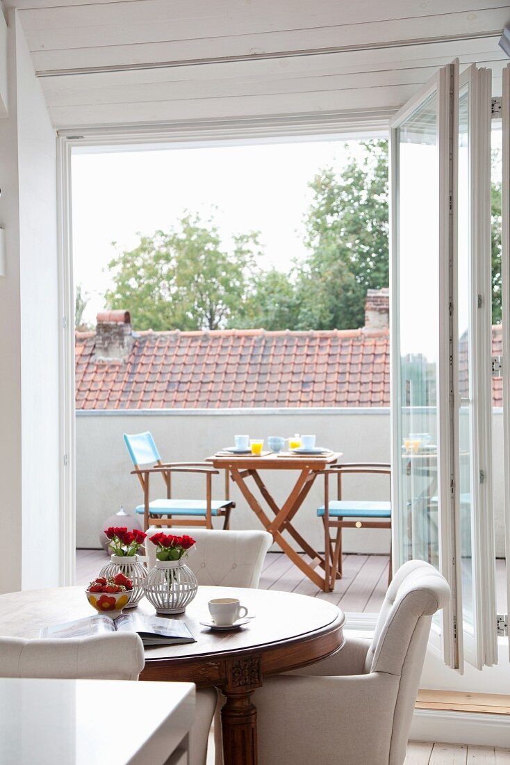 Antiker Tisch und weiße Polsterstühle vor offener Falttür und Blick auf Balkon mit Tisch und Stühlen