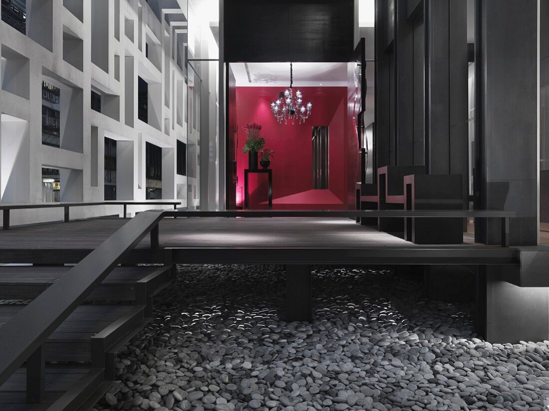 Empore mit Treppe aus dunklem Holz auf Kieselsteinbelag und Blick in offenen Vorraum auf Raumteiler und rote Wand