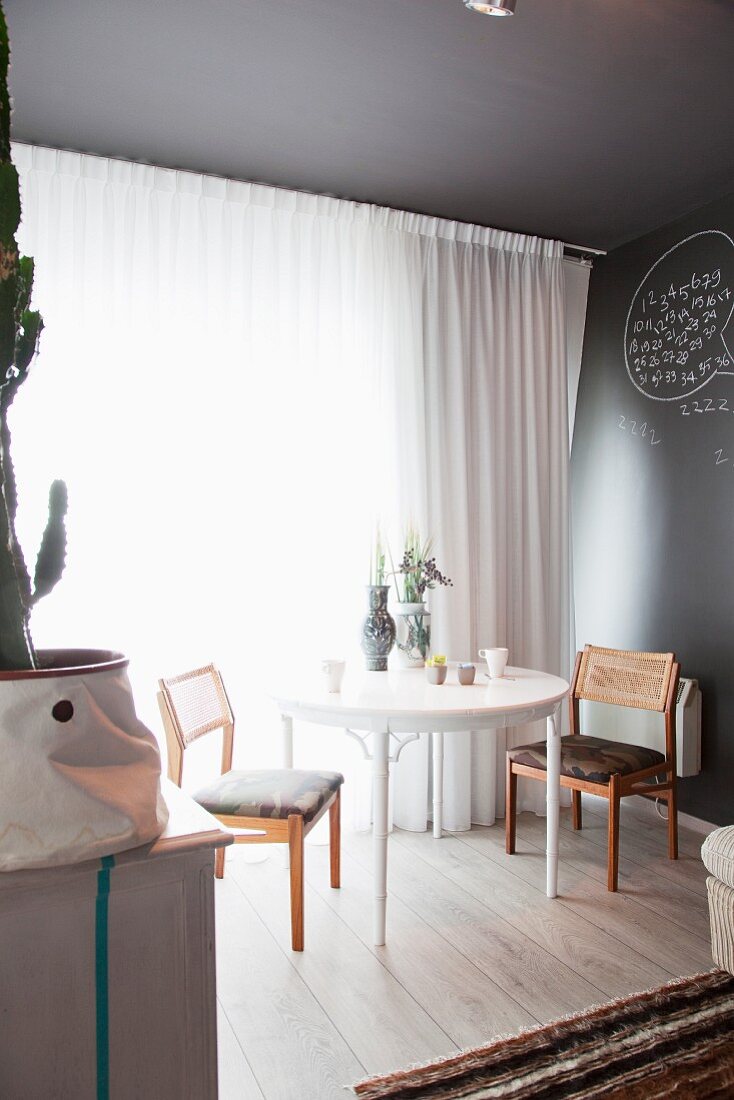 Weisser runder Tisch und Stühle vor Fenster mit luftigem Vorhang in grau getöntem Wohnraum
