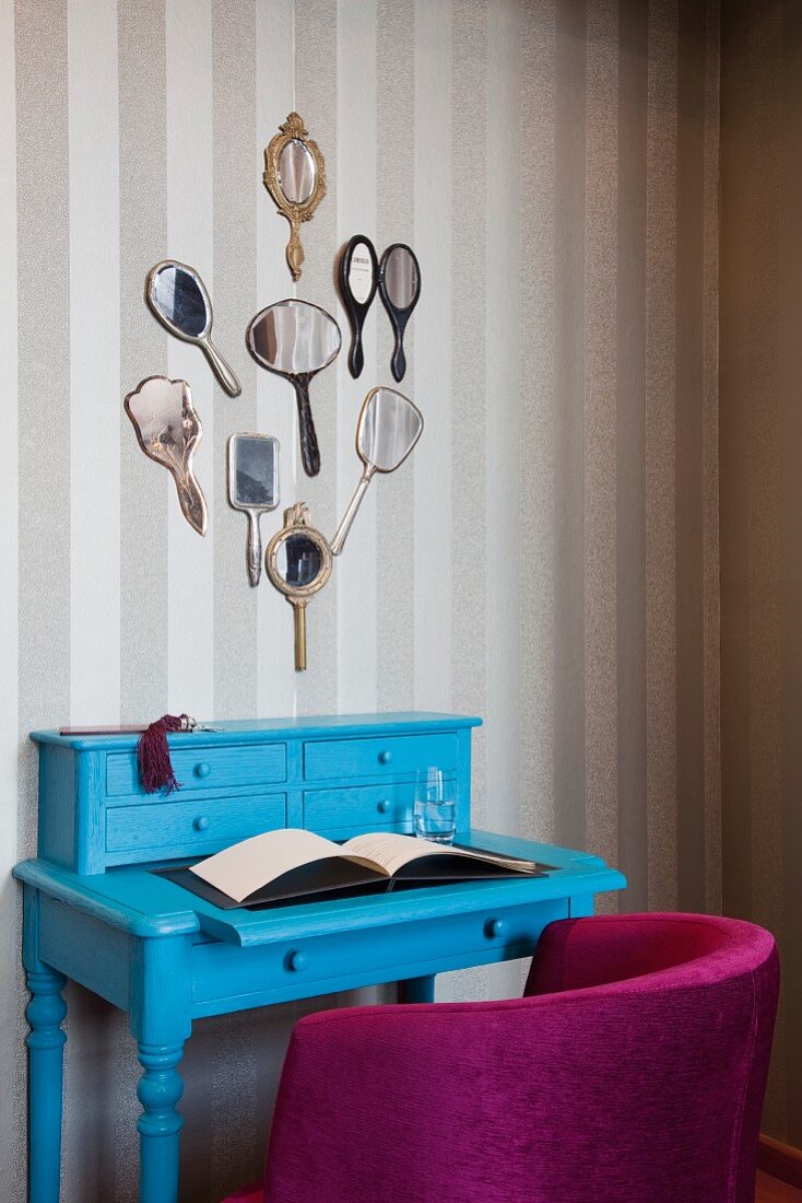 Pink armchair and azure blue dressing table below collection of hand mirrors on wall