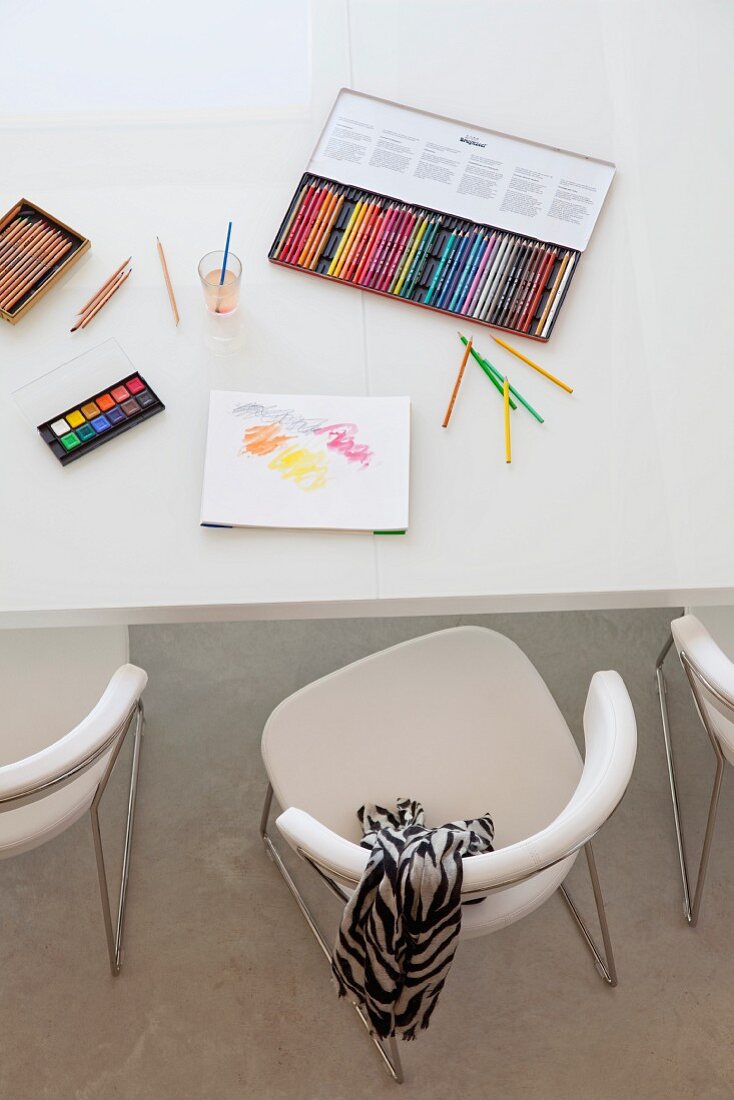 Grosse Schachtel mit Buntstiften, Wasserfarben und Zeichenblock auf weißem Tisch mit Designer Polsterstühlen