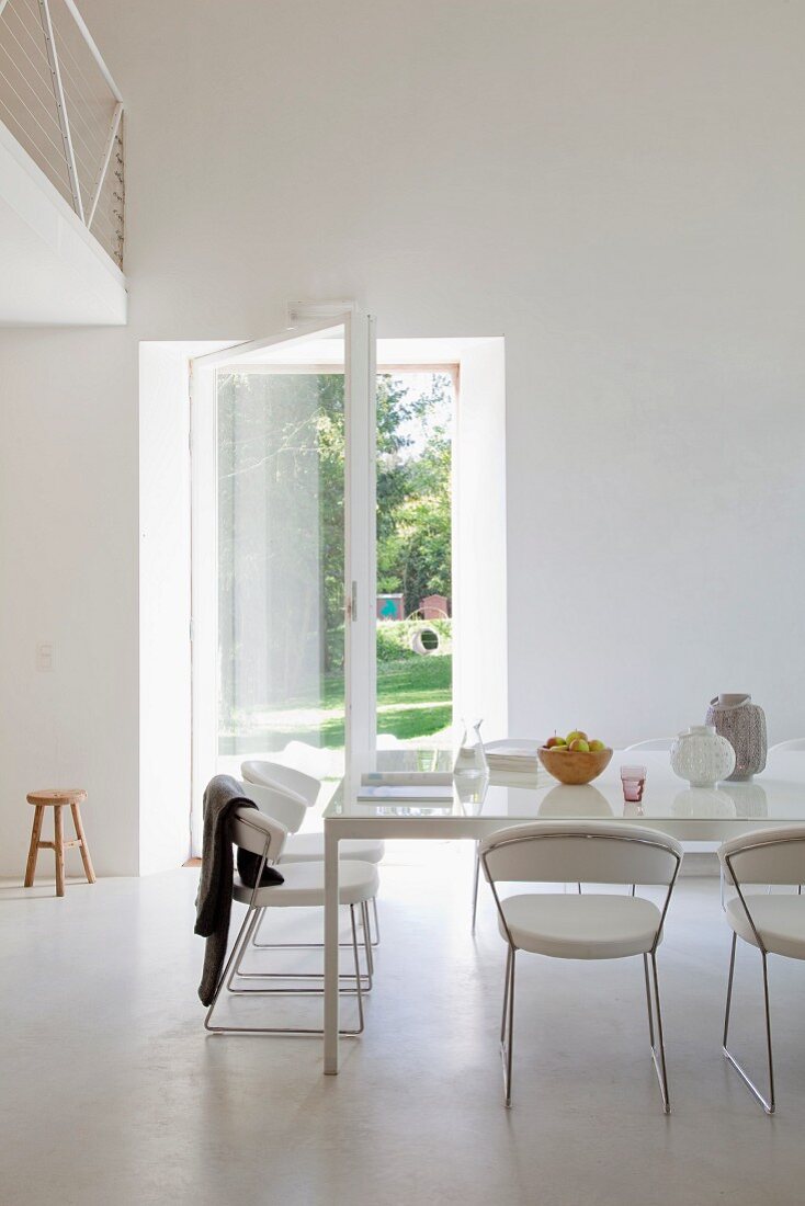 Purist common room with open door leading to garden - white leather and metal designer chairs around large table. White interior contrasting with view into green garden.