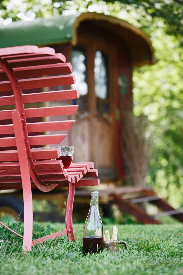 Bottle and glass of drink on lawn next to red wooden bench with old circus caravan in background
