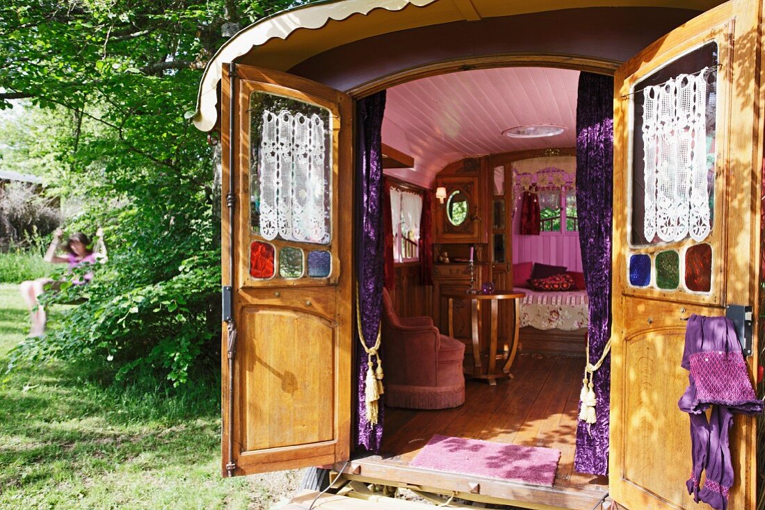 Blick vom Garten in Zirkuswagen mit offenen Türen auf rosa Sessel und rosa gestrichene Decke des holzverkleideten Innenraumes